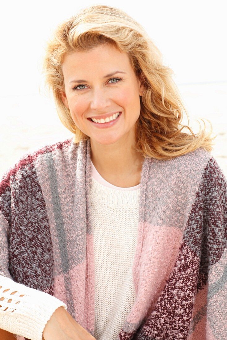 A blonde woman on a beach wearing a pink-toned shawl over her shoulders