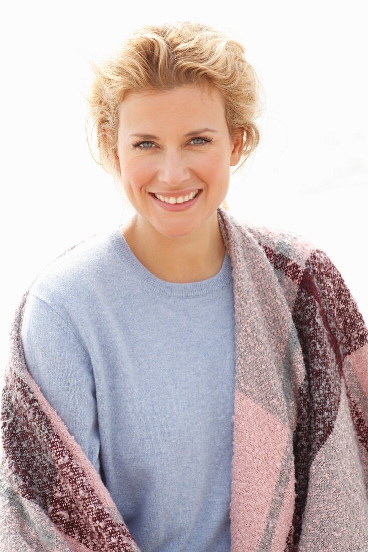 A blonde woman wearing a blue jumper and a pink-toned shawl over her shoulders