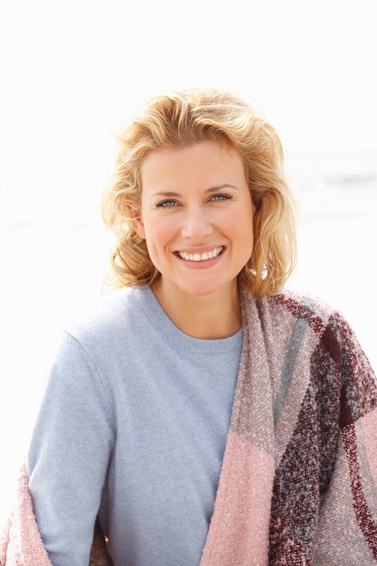 A blonde woman on a beach wearing a blue jumper and a pink-toned shawl over her shoulders