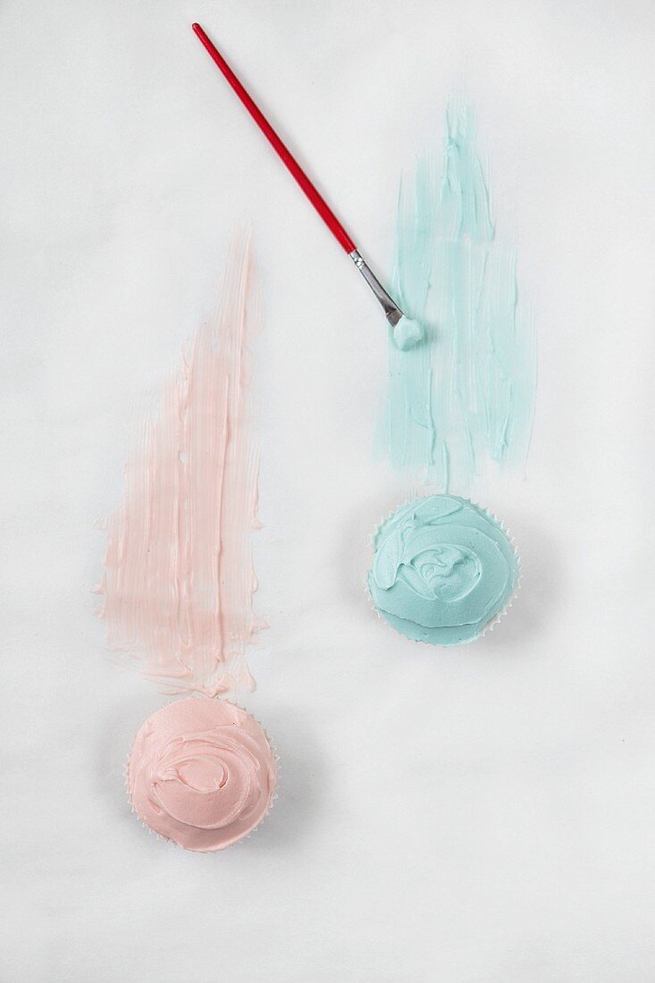 Two cupcakes with pink and blue frosting and a brush