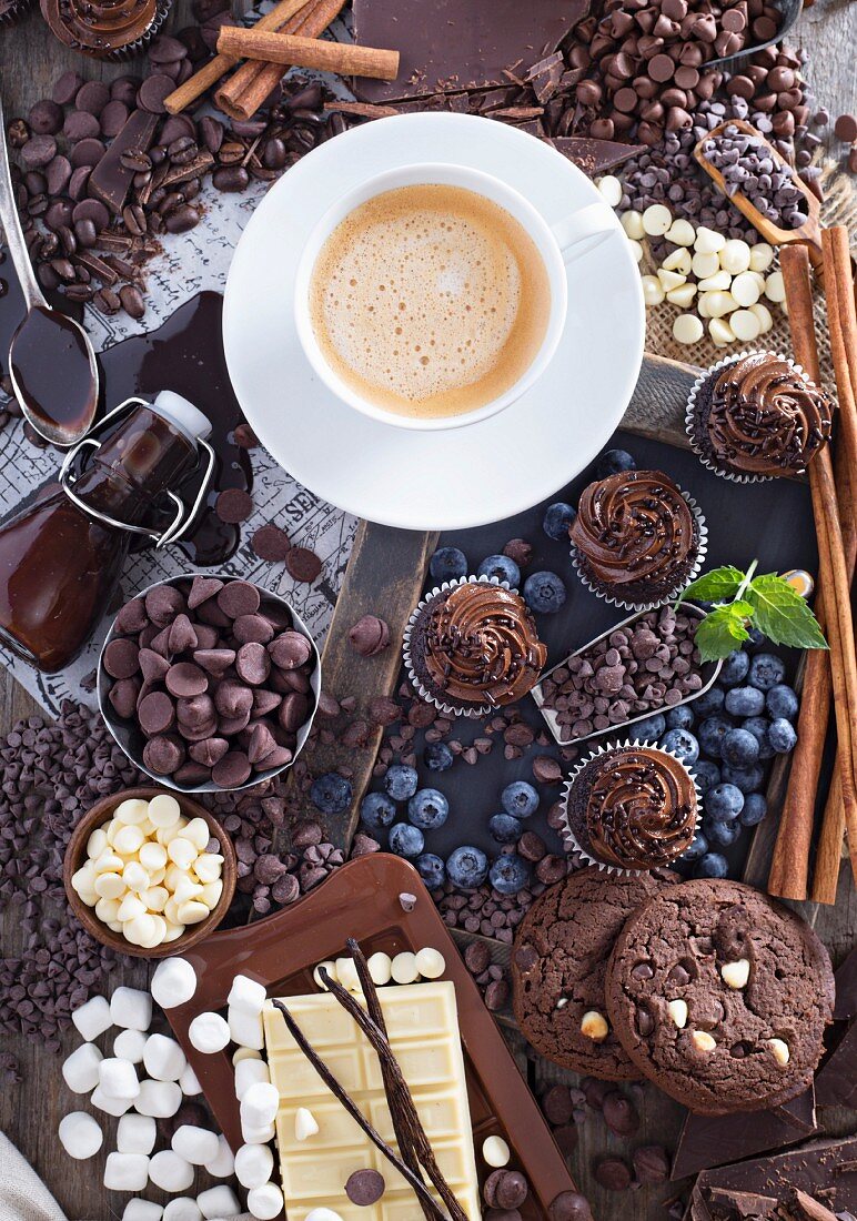Schokoladenstilleben mit Cookies, Cupcakes, Chocolatechips, Sirup, Marshmallows, Heidelbeeren, Zimtstangen und Kaffee