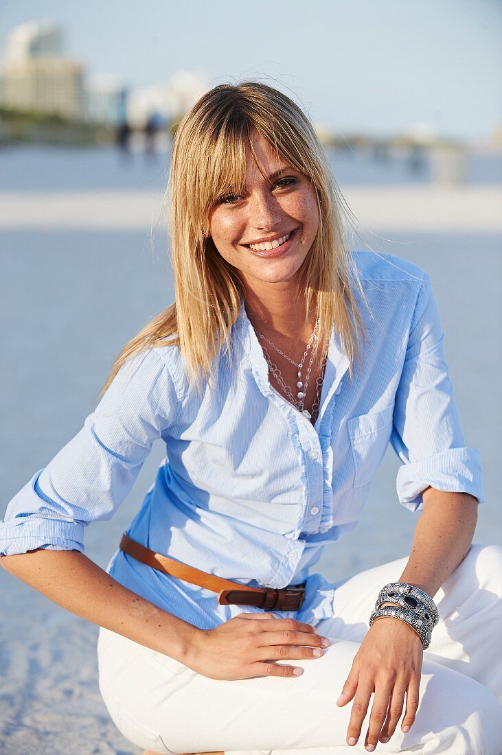 A young blonde woman on a beach wearing a light-blue shirt and white trousers