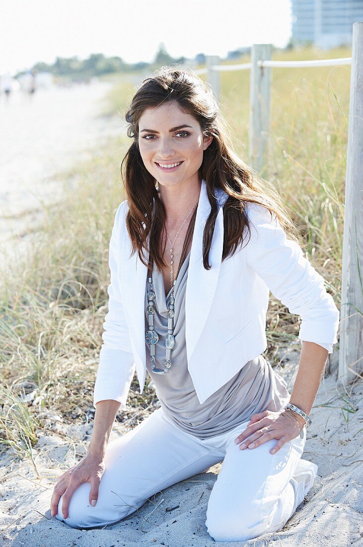 A brown-haired woman on a beach wearing a short, white jacket, a top and white trousers