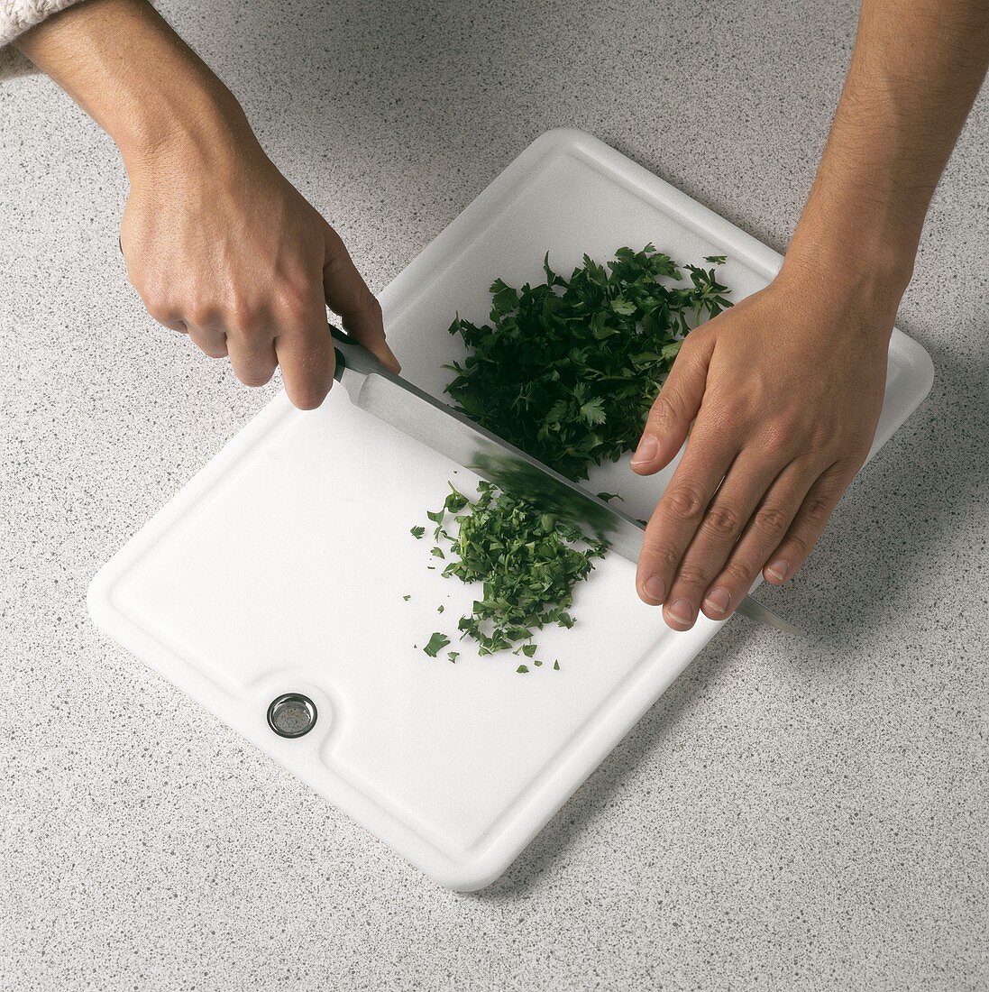 Chopping flat leaf parsley with knife on white chopping board