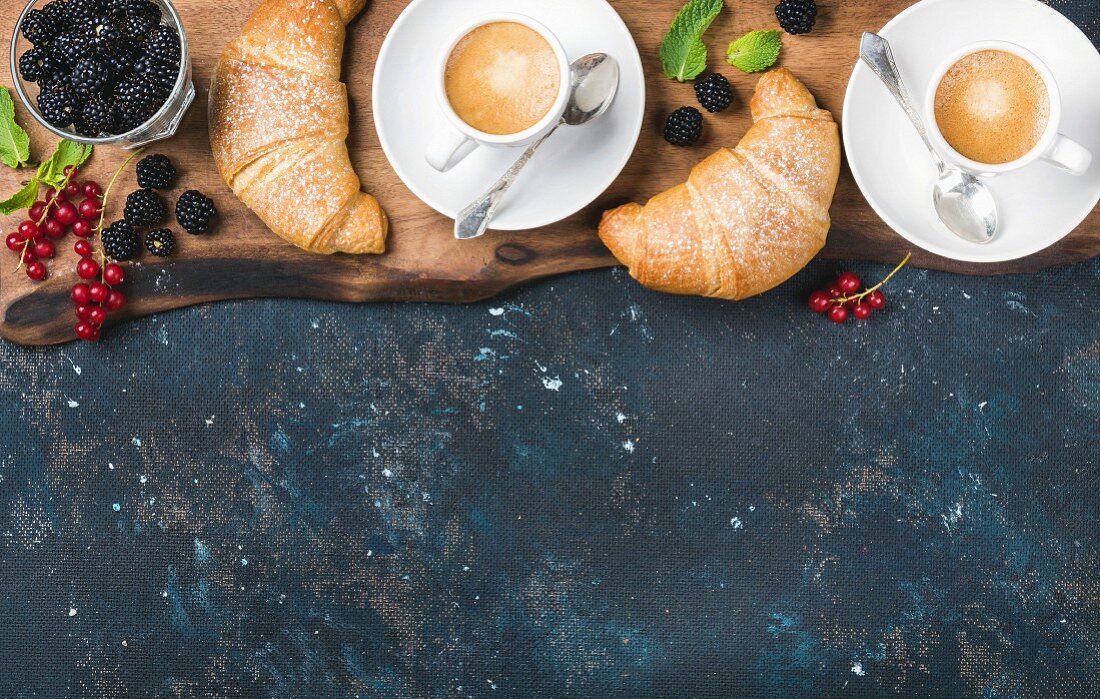 Freshly baked croissants with garden berries and coffee cups on rustic wooden board
