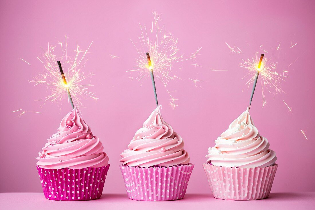 Drei rosa Cupcakes mit Wunderkerzen vor rosa Hintergrund