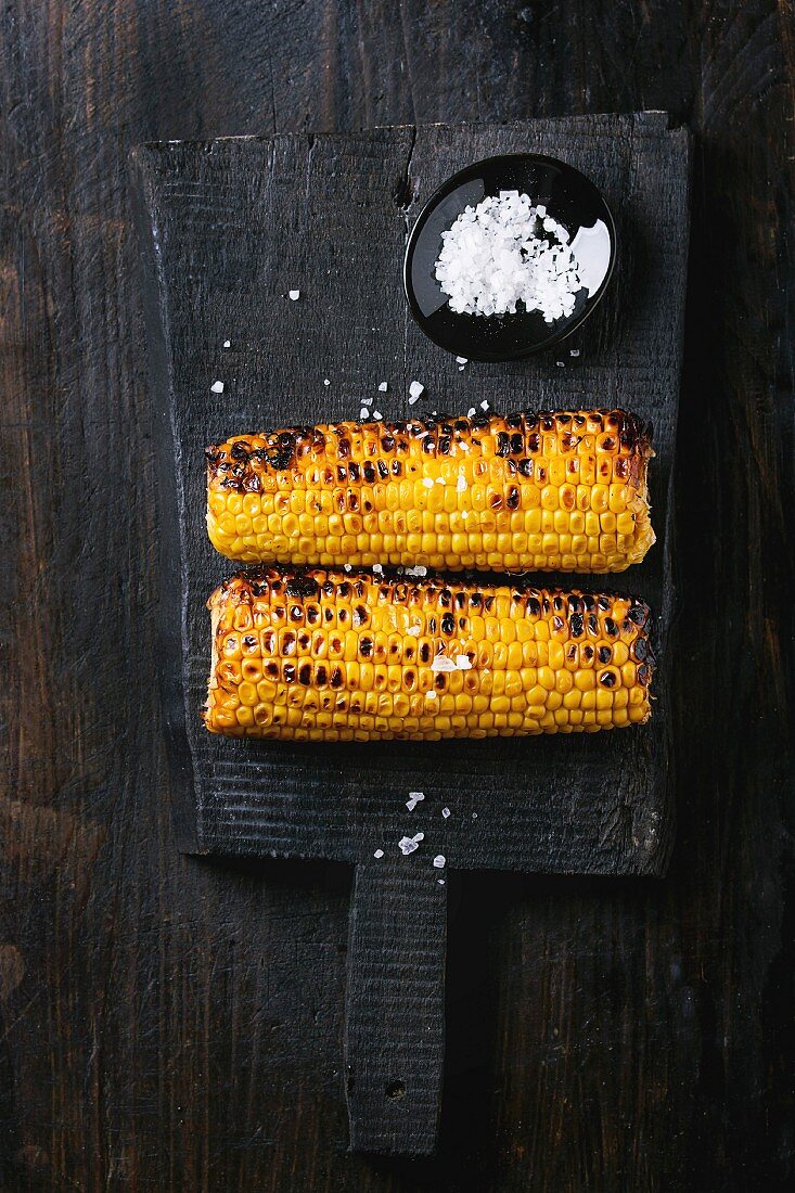 Two grilled bbq sweet corn cobs with sea salt, served on black wood chopping board