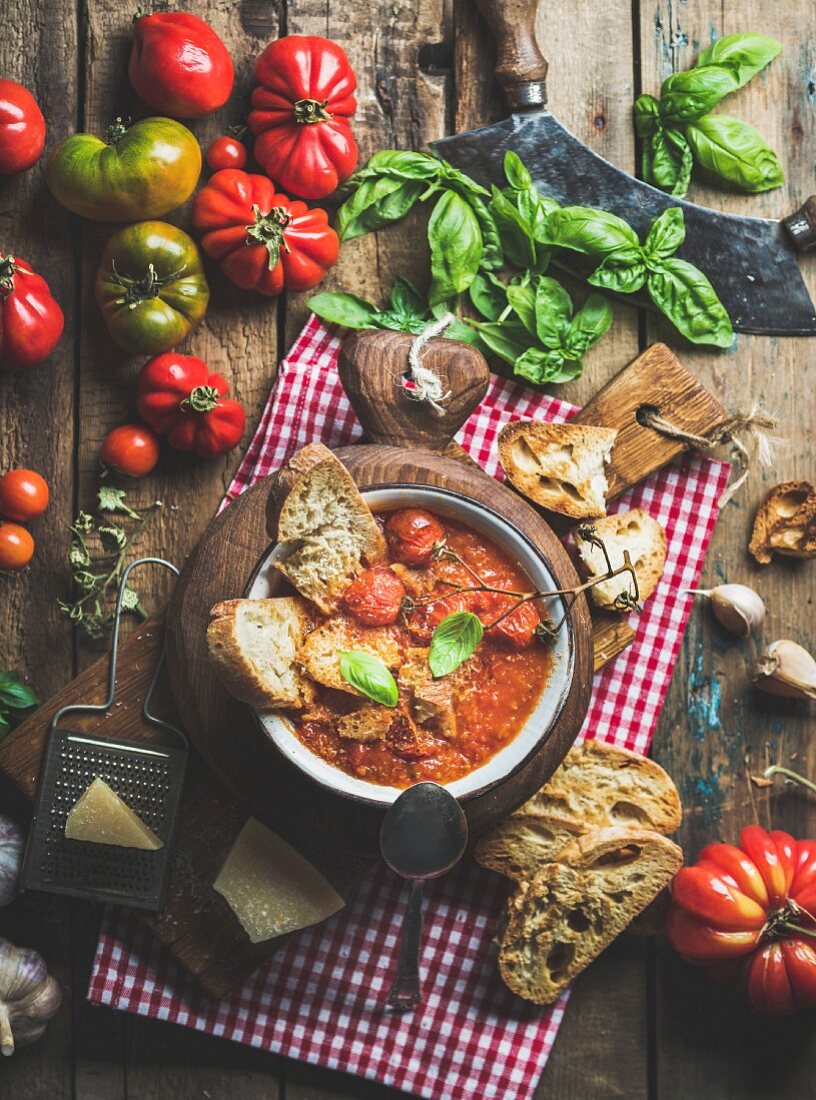 Italienische Tomaten- Knoblauch-Suppe aus gerösteten Tomaten in einer Schale auf Holzbrett mit Basilikum, Brot und Parmesan