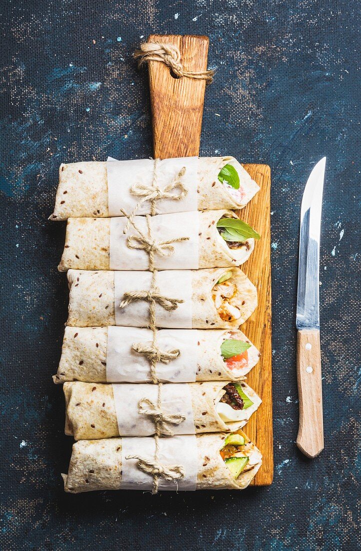 Tortilla wraps with various fillings on wooden serving board and knife over dark blue plywood background