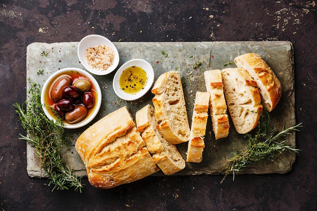 Ciabatta in Scheiben, Oliven, Olivenöl, Kräuter und Salz auf Steinplatte
