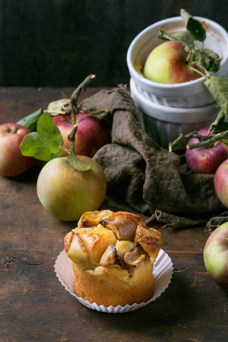 Apfel-Hüttenkäse-Minikuchen in Papierförmchen vor frischen Äpfeln auf dunklem Holzhintergrund