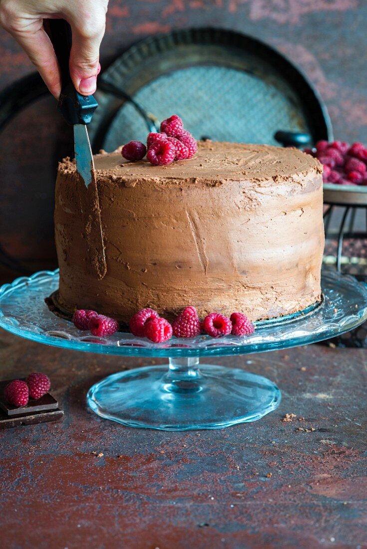 A hand using a spatula to smooth the chocolate cream on the side of a chocolate cake with ganache and raspberries