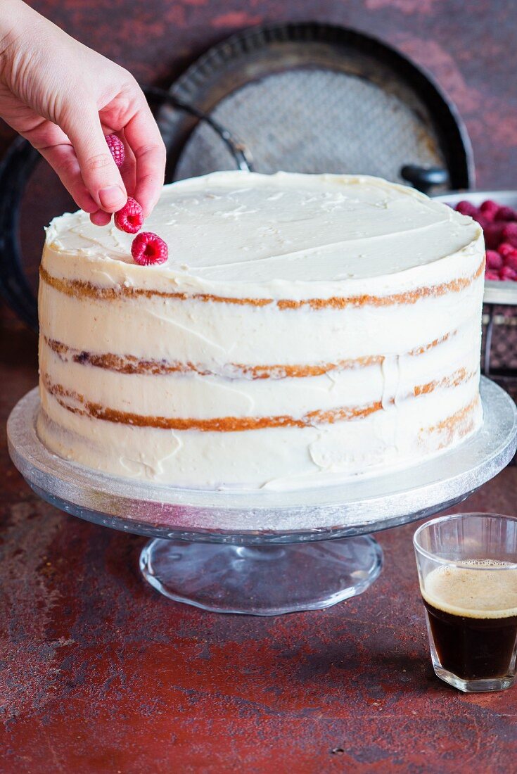 A vanilla layer cake being decorated with raspberries