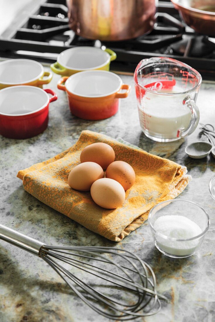 Eier, Milch und Zucker mit Schneebesen und Backförmchen neben Gasherd