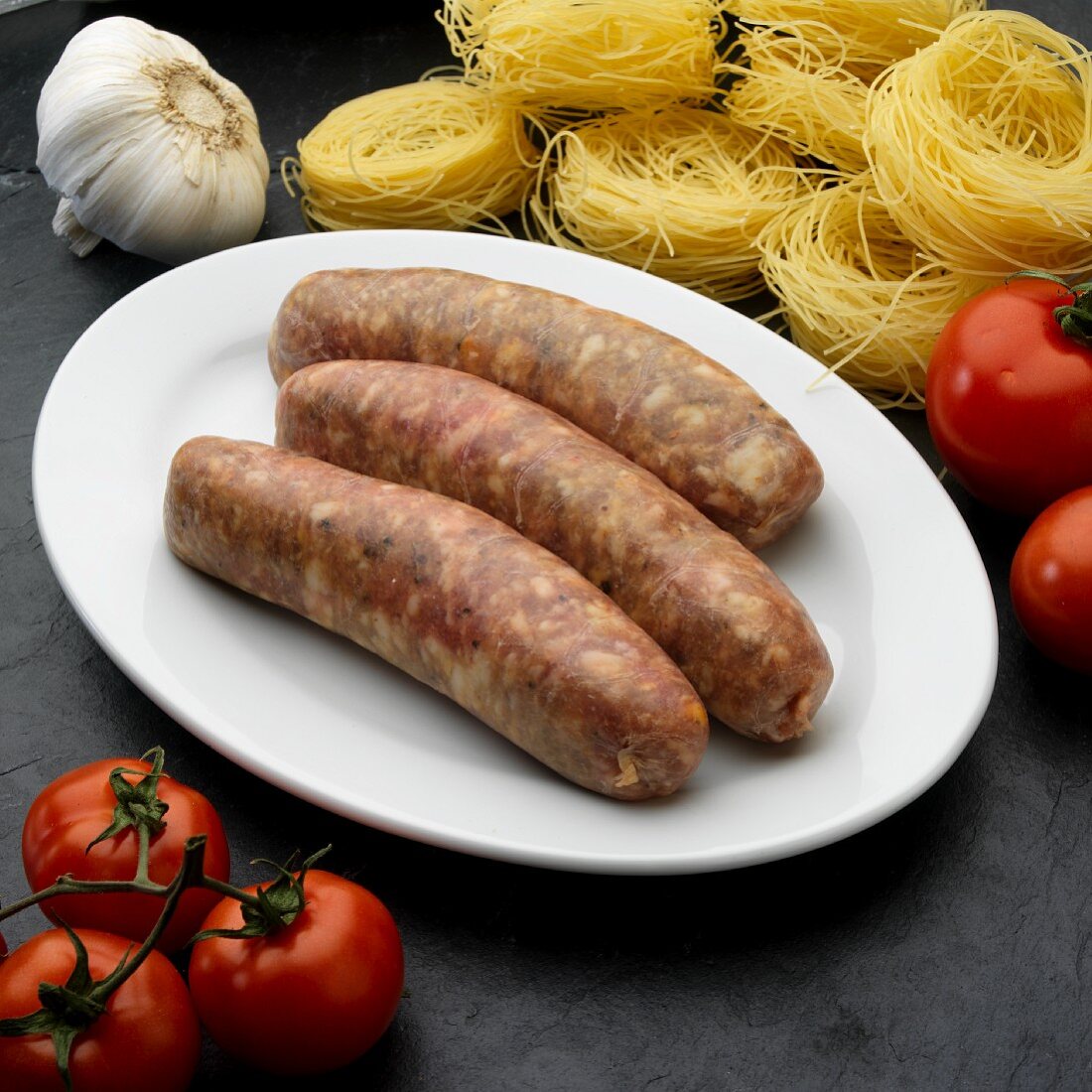 Drei frische italienische Bratwürste (Salsiccia) auf Teller umgeben von Zutaten