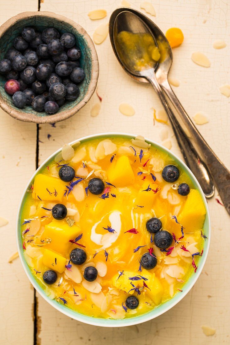 Smoothie Bowl mit Mango, Ananas, Joghurt, Heidelbeeren, Mandelblättchen und Blüten