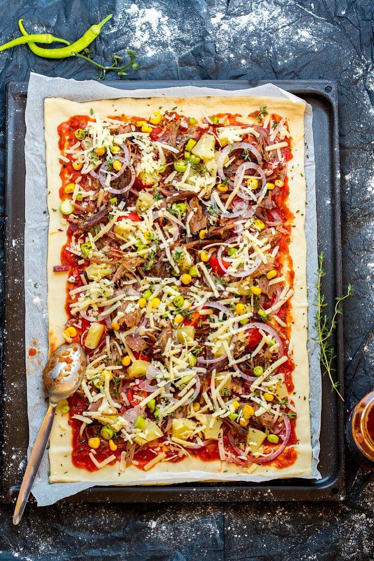 An unbaked pizza with pulled pork, sweetcorn, pineapple and onion on a baking sheet