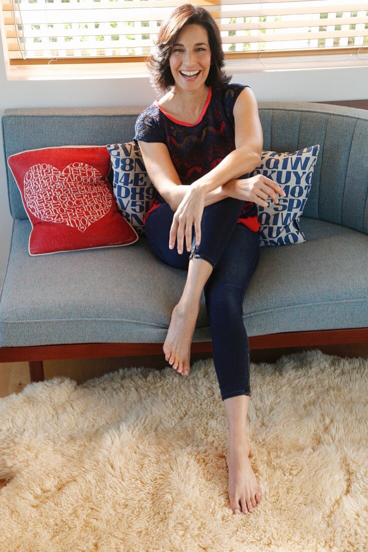 A brunette woman wearing a lace top and leggings and sitting on a grey sofa