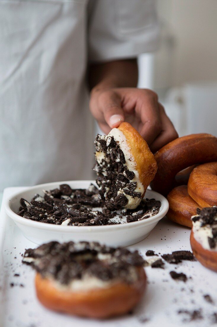 Doughnuts with white chocolate icing being dipped in crumbled Oreo biscuits