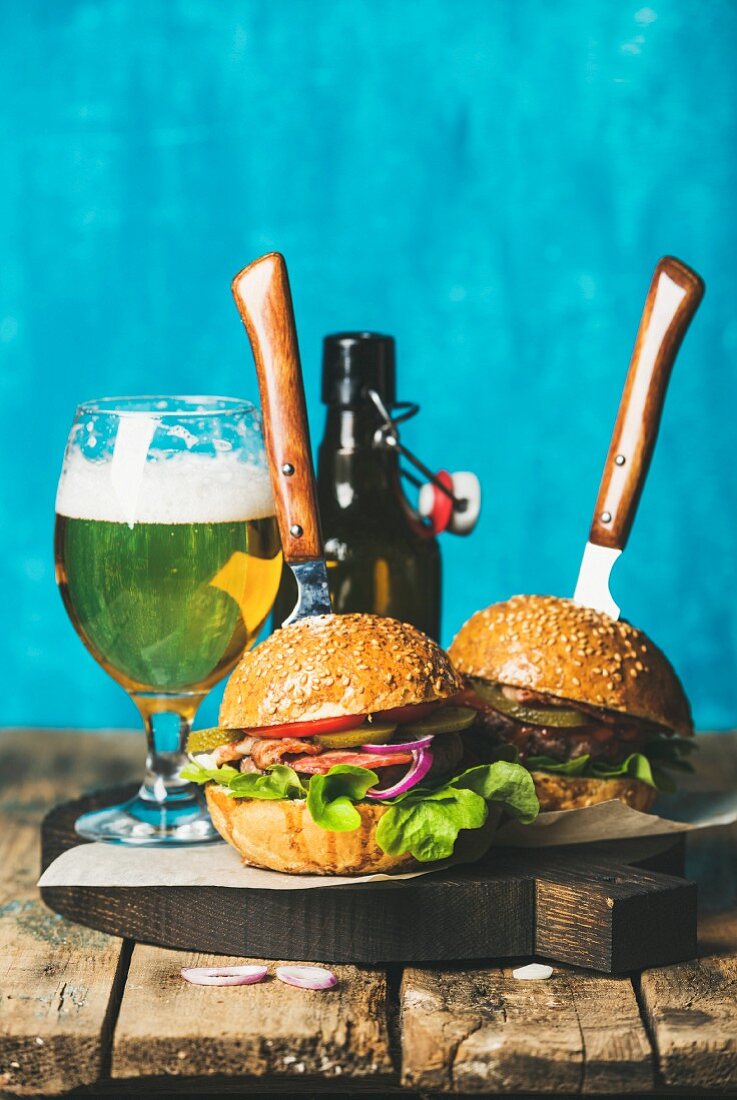 Two homemade beef burgers with crispy bacon, onion, pickles, vegetables, glass and bottle of wheat beer