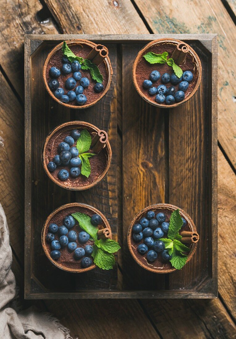 Homemade Tiramisu dessert in glasses with cinnamon sticks, mint leaves and fresh blueberries in wooden tray