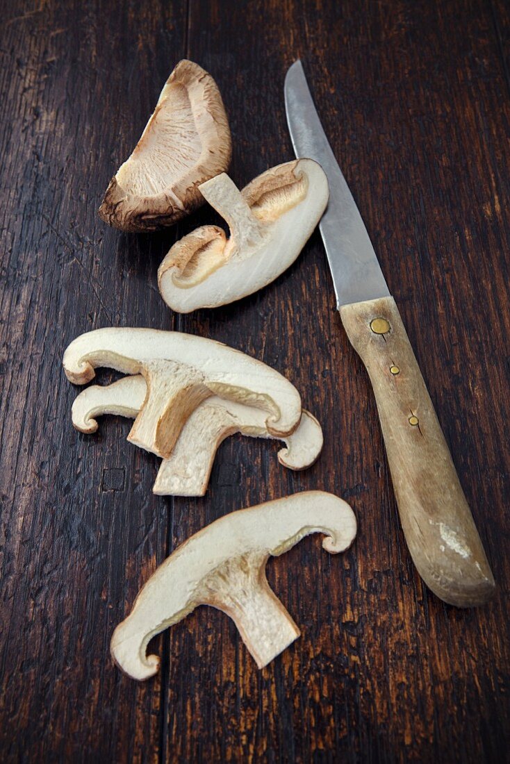 Ein Shiitakepilz, in Scheiben geschnitten, mit Messer auf Holzuntergrund