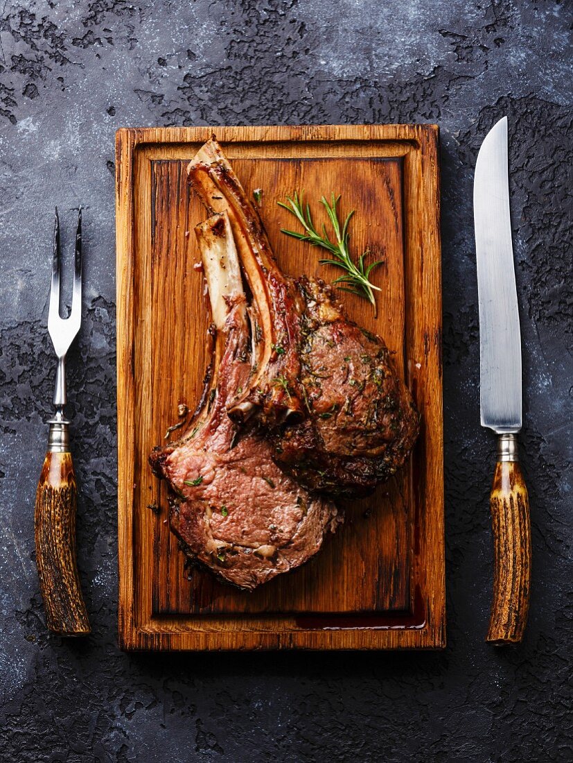 Roasted beef ribs on bone on wooden cutting board with knife and fork carving set on dark background