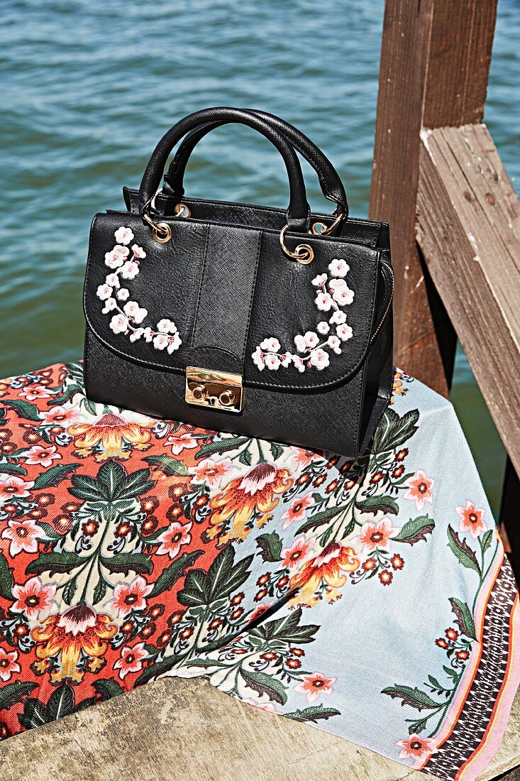 A black handbag with an embroidered flower pattern and a scarf with a flower pattern on a wooden jetty