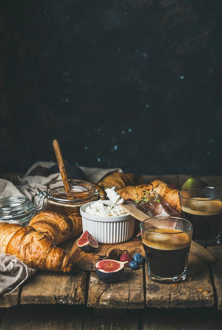 Frühstück mit Croissants, Ricotta, Feigen, Beeren, Schinken, Honig und Espresso auf rustikalen Holztisch