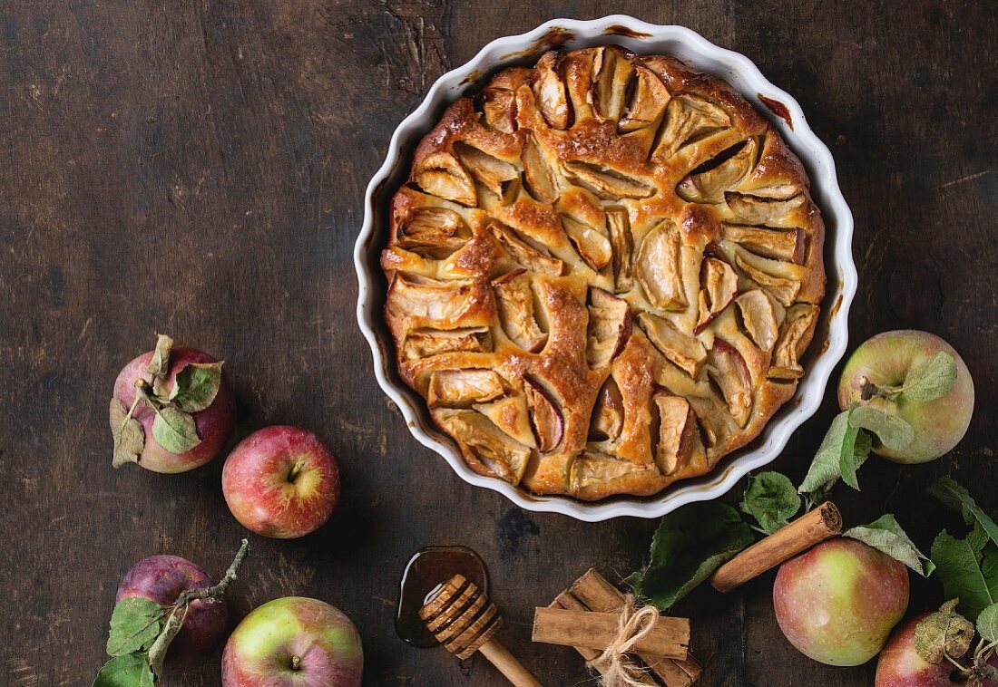 Apple cake pie in a white ceramic flan dish with fresh apples, honey and cinnamon sticks