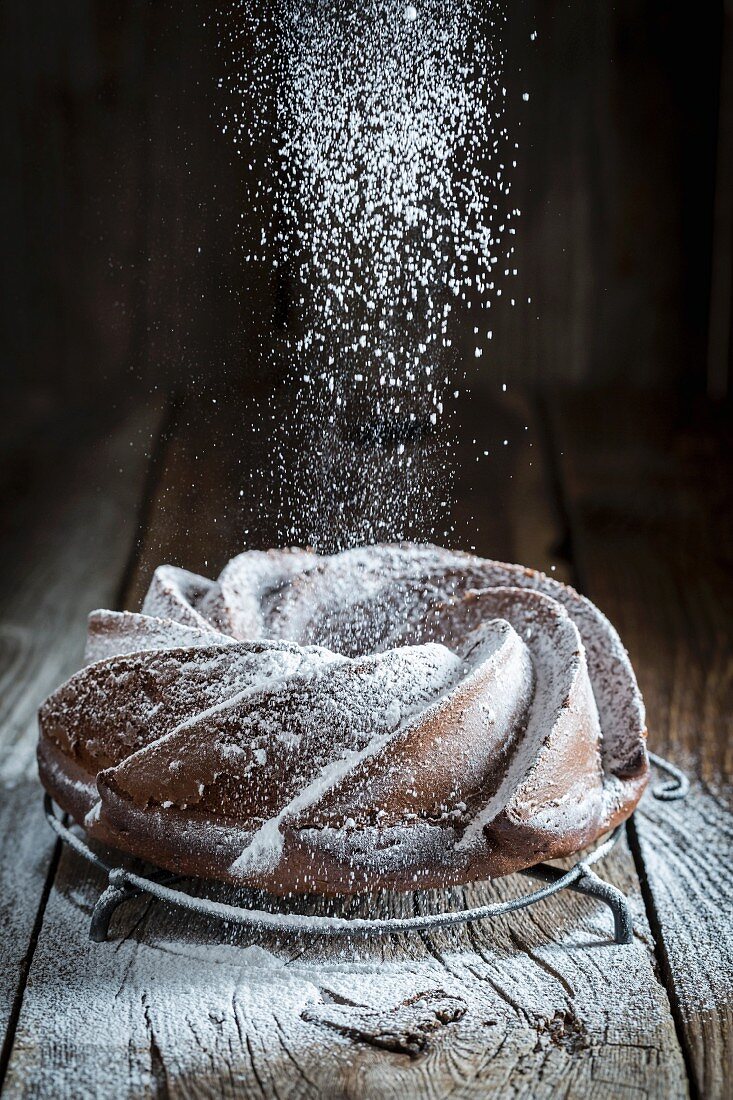 A Bundt cake on a wooden table being dusted with icing sugar
