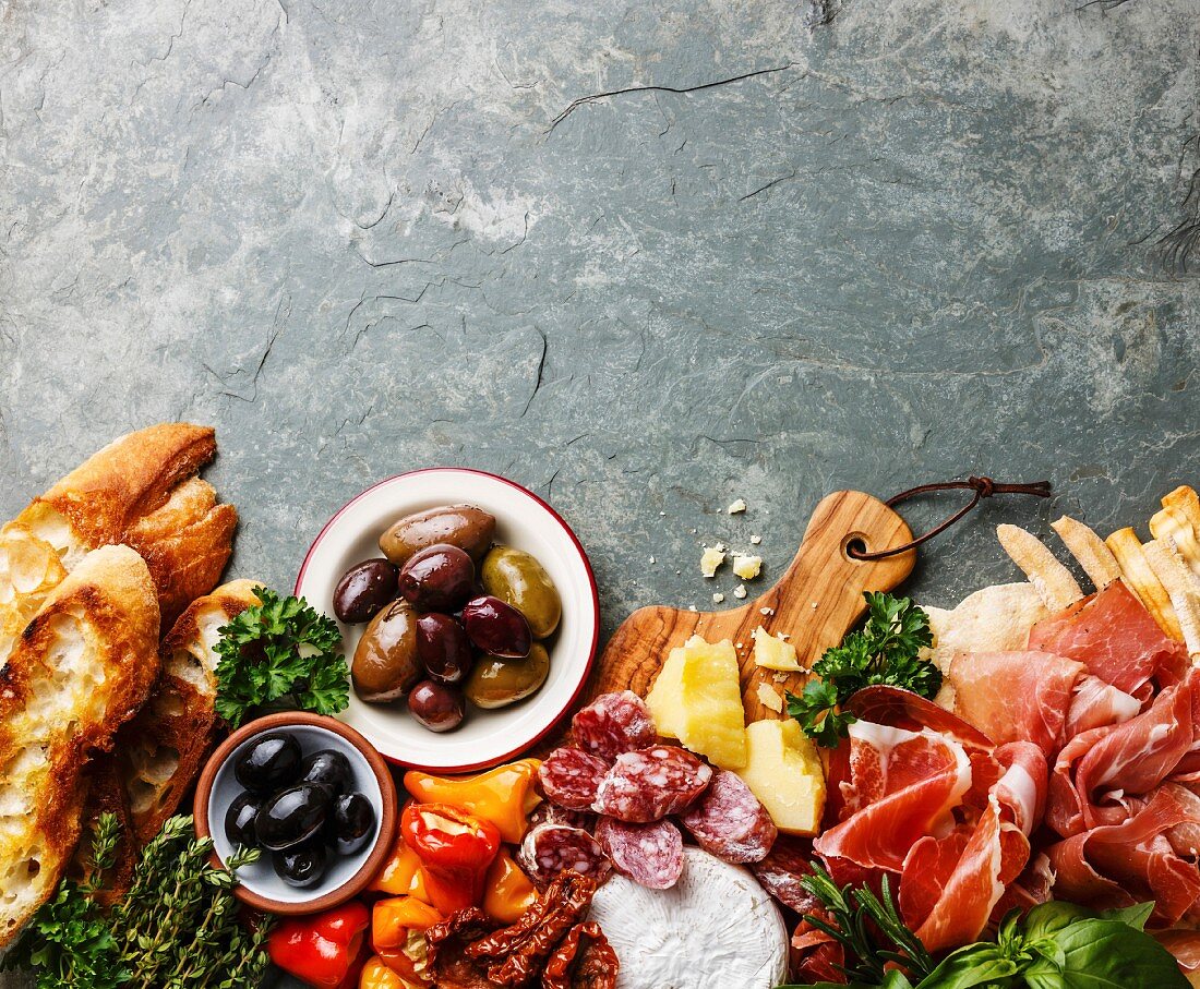 Italian food ingredients background with ham, salami, parmesan, olives, bread sticks on stone slate