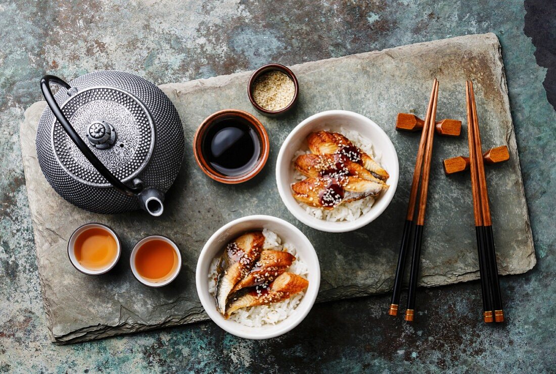 Eel with sauce and sesame seeds on a bed of rice with chopsticks, a teapot and tea bowls