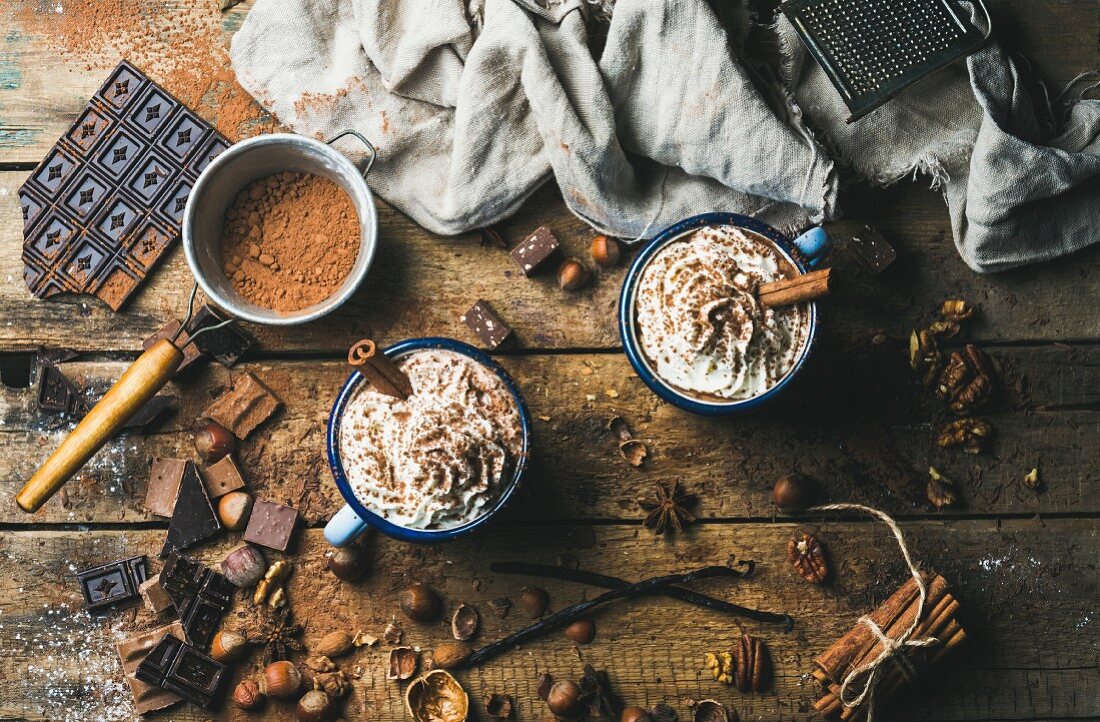Heiße Schokolade mit Sahne, Nüssen und Zimt in Emaille-Becher und Zutaten auf rustikalem Holzuntergrund