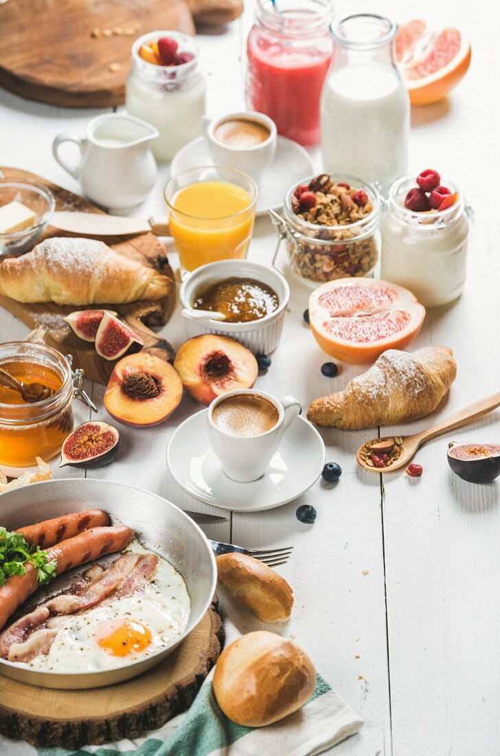 Fried egg with sausages and bacon, bread, croissants, jam, butter, fruits, smoothie, orange juice, yogurt, granola, milk and coffee