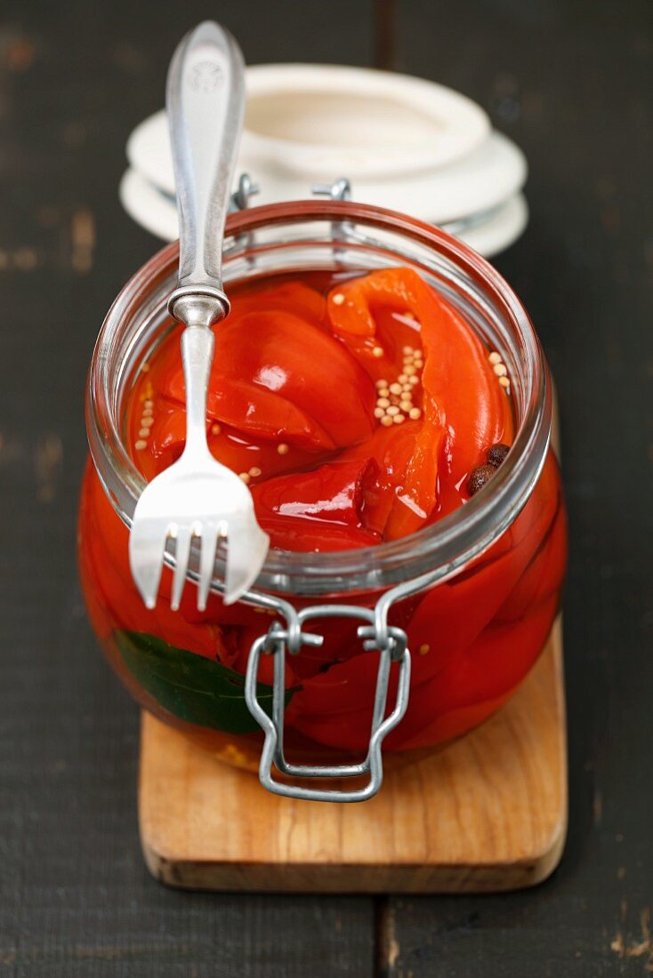 Pickled peppers in a glass jar with a fork on a chopping board