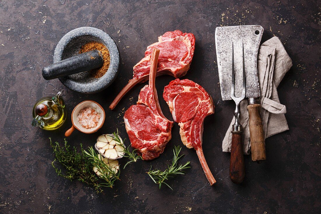 Raw veal chops with herbs and spices and vintage kitchen utensils on a dark surface
