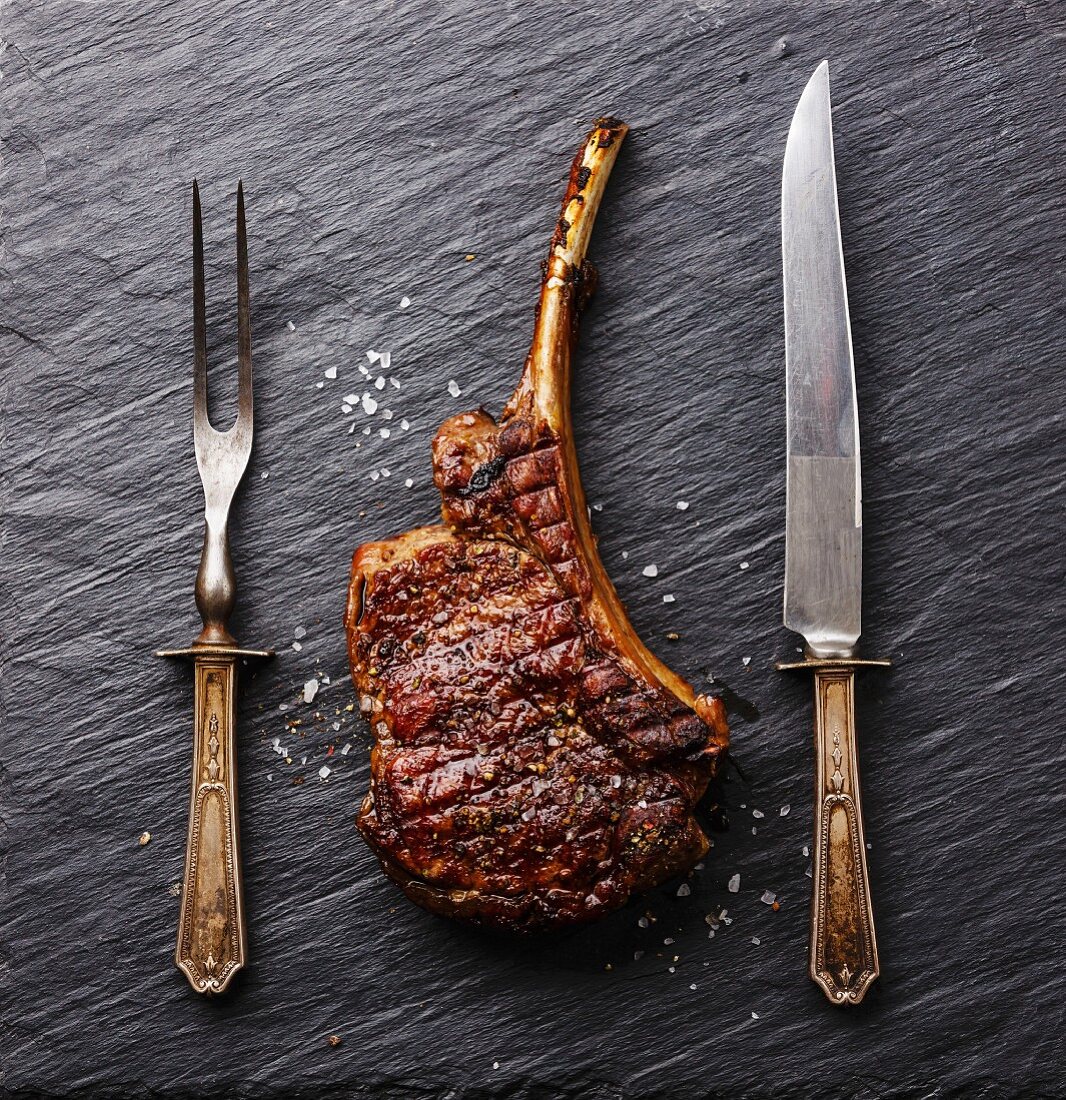A roasted beef rib steak with a knife and fork on a slate surface