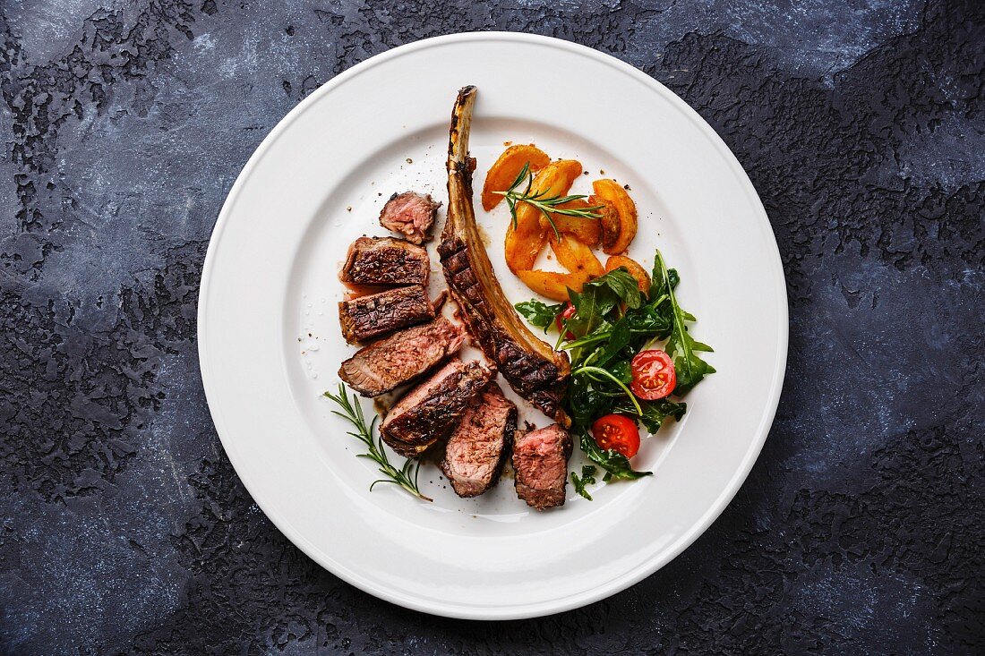 A grilled beef rib steak on a white plate with potato wedges and tomato and rocket salad