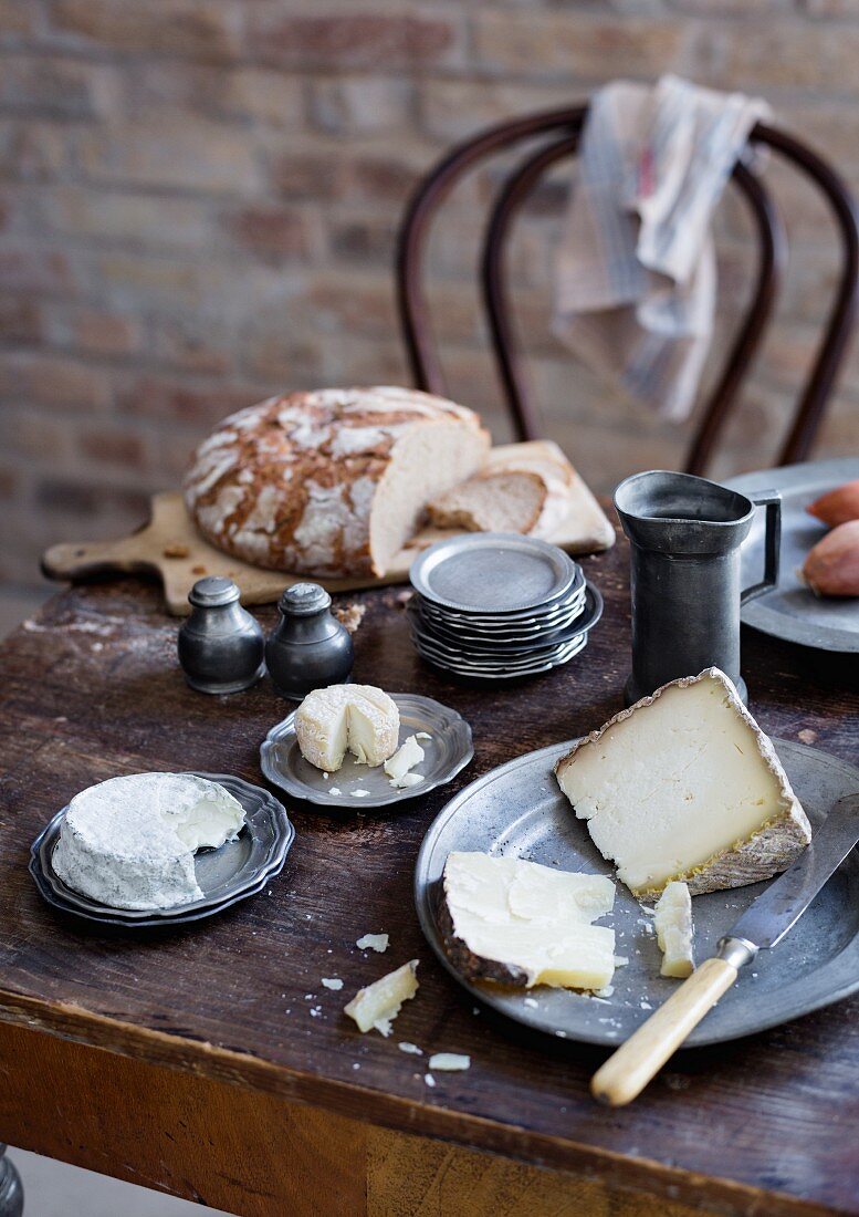 Rustikal gedeckter Tisch mit Zinngeschirr, Käse und Brot vor Backsteinwand