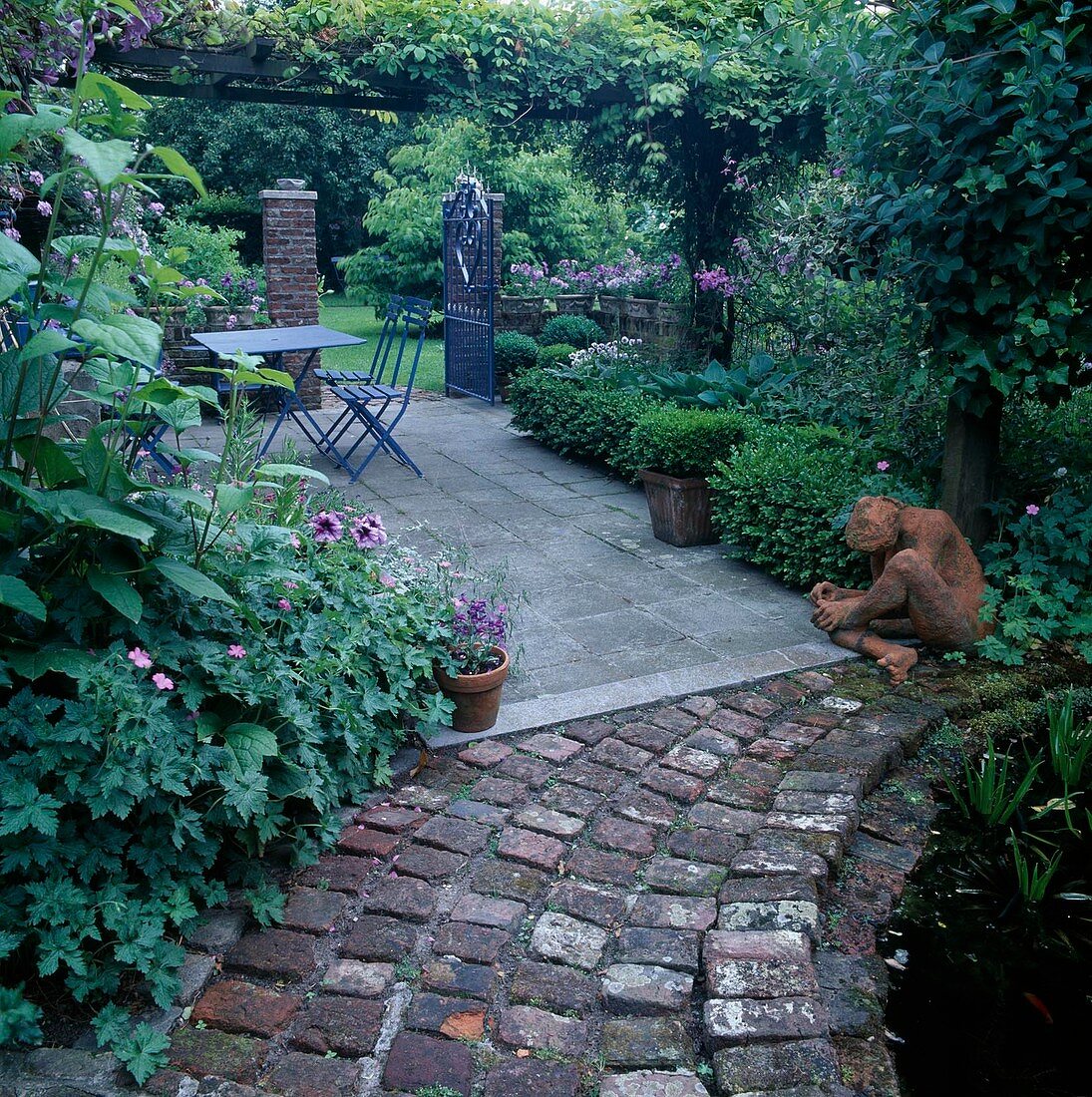 Shady terrace under pergola, blue seating group, garden gate, Buxus (boxwood), terracotta figure