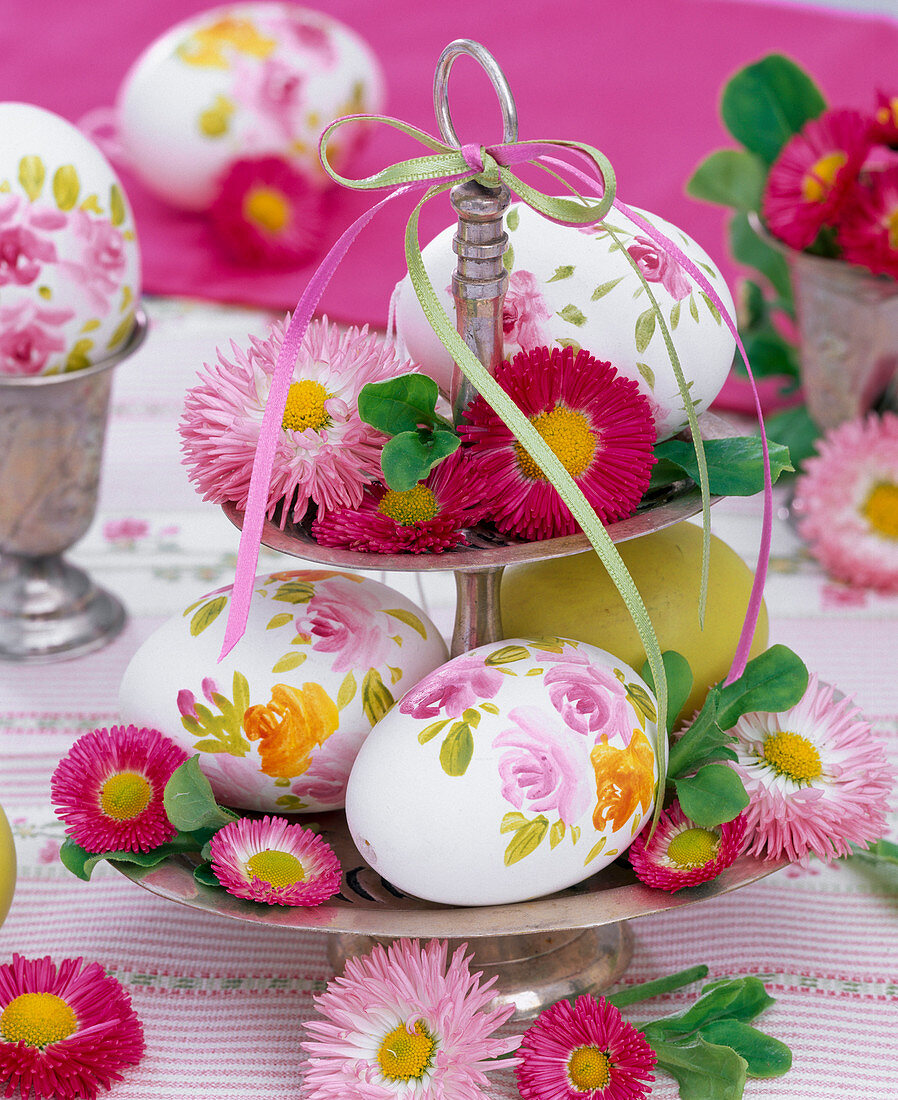 Bellis (Gänseblümchen) und bemalte Eier auf silberner Etagere mit Bändern