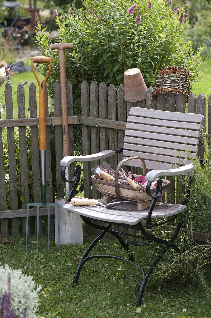 Korb mit Kleingeräten auf Gartenstuhl, Spaten und Grabgabel am Zaun