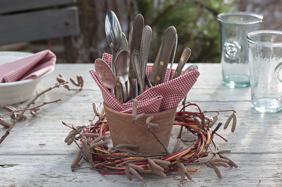 Cutlery with red and white checked napkin in terracotta vase, wreath of Corylus