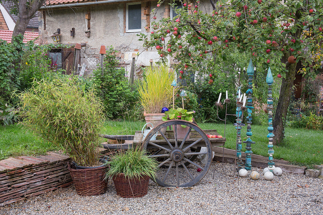 Gravel terrace with small dry stone wall made of old roof tiles, old barrow with wooden wheels, Fargesia murielae (bamboo), grasses, decorative plugs made of handmade ceramics, candlesticks, baubles, apple tree (Malus)
