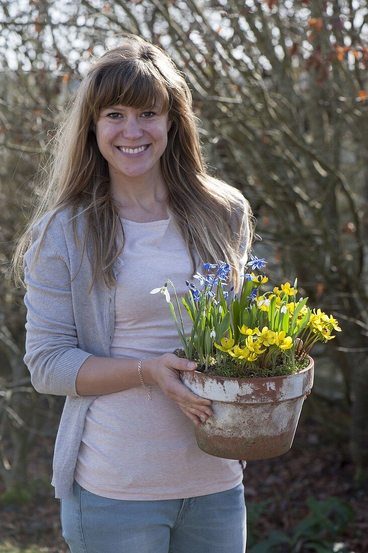 Frau bringt Tontopf mit Scilla (Blausternchen), Eranthis (Winterlingen)