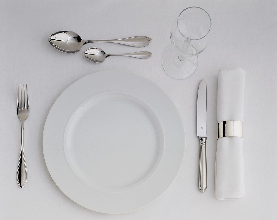 A place setting with white plate, cutlery, napkin & wine glass