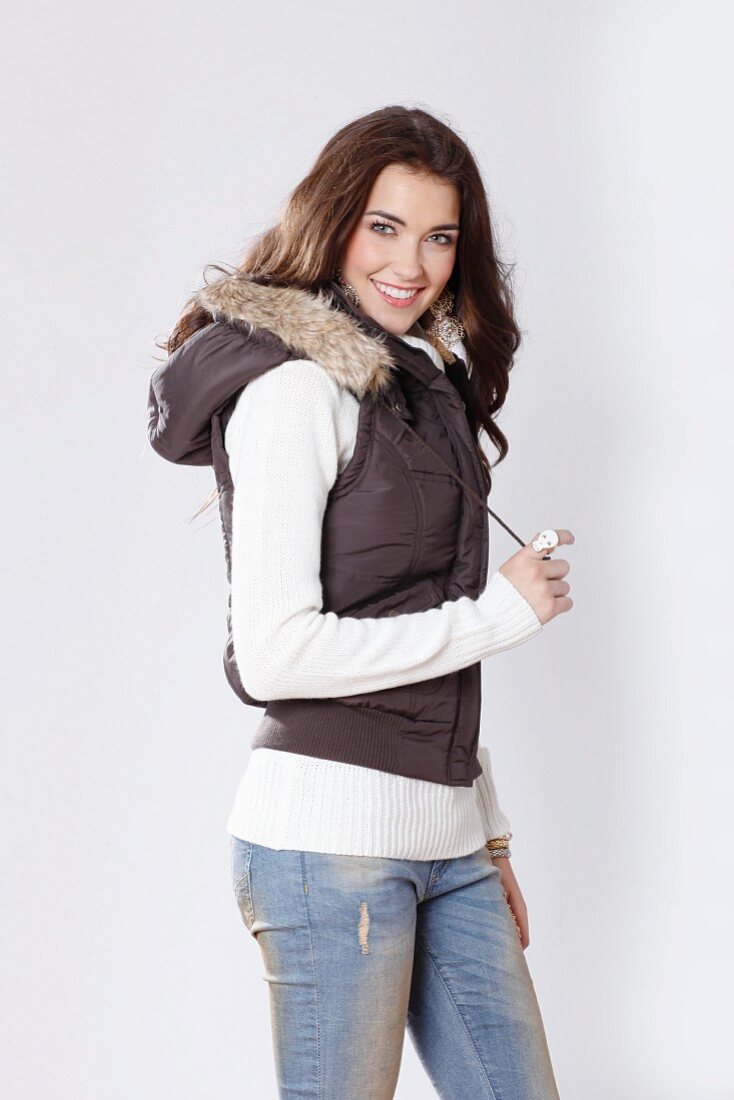 A brunette woman wearing a white knitted jumper, a brown bodywarmer with a fur-trimmed hood and jeans