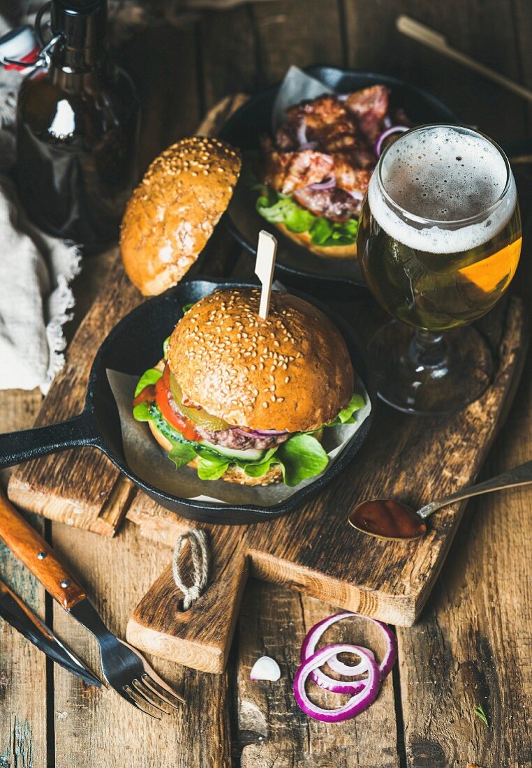 Homemade beef burgers with crispy bacon and vegetables, glass of wheat beer on rustic serving board