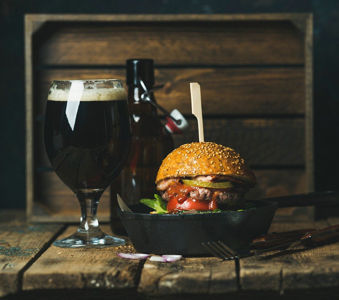 Homemade beef burger with crispy bacon, fresh vegetables and ketchup in black cast iron pan, with glass of dark beer
